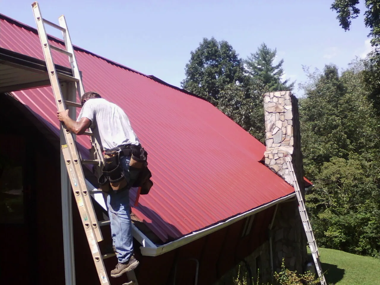 Roofer on a ladder beside a red metal roof and stone chimney — Soffit & Fascia Repair by Roofing Brothers in Murrysville